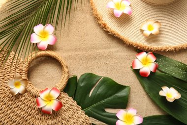 Farklı tropik yapraklara sahip güzel tesisat çiçekleri, hasır çanta ve kumlu arka planda şık bir şapka.