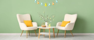 Oturma odasının içinde beyaz koltuklar, masa ve Paskalya süslemeleri yeşil duvarın yanında.