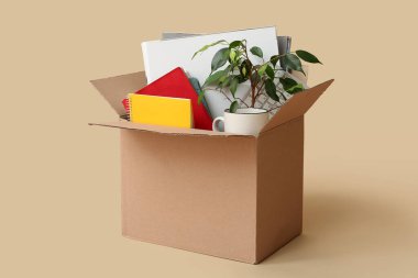 Ofis malzemelerinin olduğu karton taşınma kutusu, bej arka planda fincan ve bitki.