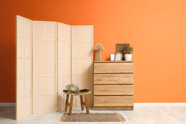 Oturma odasının içinde çekmeceler, masa ve turuncu duvarın yanında katlanan ekran var.