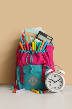 Turuncu duvarın yanındaki beyaz masanın üzerinde farklı kırtasiye malzemesi ve alarm saati olan renkli okul çantası.