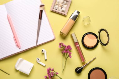 Makyaj malzemeleri, modern kulaklıklar, güzel çiçekler ve sarı arka planda not defteri.