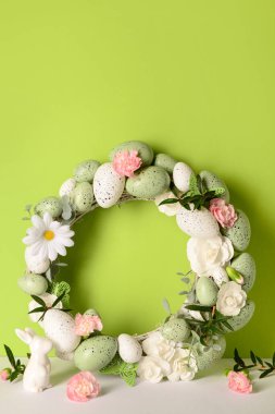 Küçük tavşan, yeşil arka planda çiçekler ve yumurtalarla süslenmiş Paskalya çelengi.