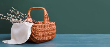 Basket with pussy willow branches and Easter eggs on wooden table against dark green background with space for text