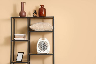 Raflarda modern elektrikli vantilatör ısıtıcısı ve bej duvarın yanında dekor var.