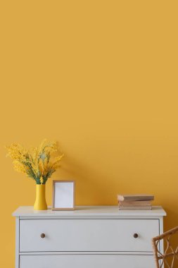 Sarı duvarın yanında vazo, çerçeve ve kitaplar olan dolap.