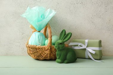 Boyanmış Paskalya yumurtaları, hediye çantası ve yeşil ahşap masada oyuncak tavşancık olan hasır sepet.