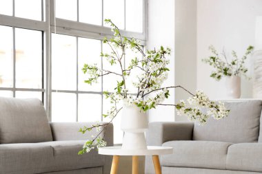 Aydınlık oturma odasındaki gri kanepenin yanındaki çekmecelerin üzerinde çiçek açan bir vazo.