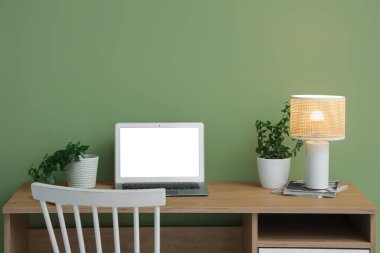Minimalist çalışma alanı, dizüstü bilgisayarı ve yeşil duvarın yanındaki ahşap masada parlayan masa lambası.