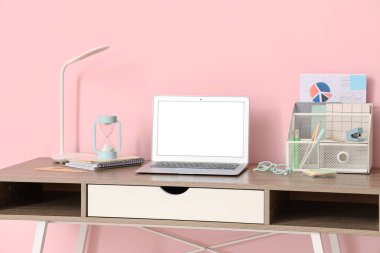 Ofisteki pembe duvarın yanında modern dizüstü bilgisayarı, kırtasiyesi ve lambası olan şık bir iş yeri.