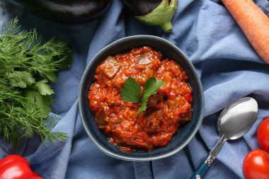 Bir kase sebze yahnisi, patlıcan, domates ve mavi peçeteli otlar.