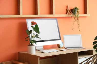 Turuncu duvarın yanında modern bilgisayar, dizüstü bilgisayar ve ev bitkileriyle rahat çalışma alanı