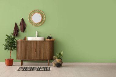 Seramik lavabo, ayna ve ahşap çekmeceler şık bir banyoda yeşil duvarın yanında.