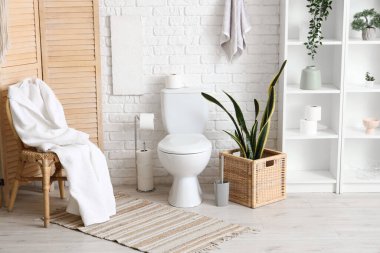 Tuvalet klozeti, bornozlu sandalye ve tuvaletteki beyaz tuğla duvarın yanında çiçek.