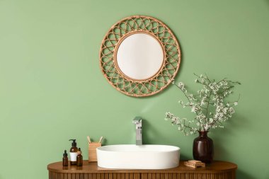 Çiçek açan dalları olan vazo, banyo malzemeleri ve banyodaki yeşil duvarın yanındaki çekmecelere batan lavabo.