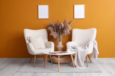 Konforlu koltuklar, masa ve vazo, oturma odasının içinde sarı duvarın yanında otlar var.