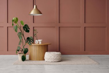 Odanın içindeki kahverengi duvarın yanında ahşap masa, puf ve çiçek var.