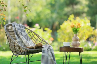 Rahat hasır koltuk, kitap, kahve fincanı ve vazo dolusu lale çiçekleri parkta, dışarıda.