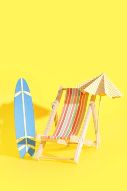 Sarı arka planda minyatür sörf tahtası, şezlonga ve şemsiyeye sahip yaratıcı kompozisyon