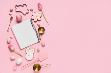 Tarifler, pişirme araçları, paskalya yumurtaları ve pembe arka planda kurabiyeler için boş defter içeren kompozisyon