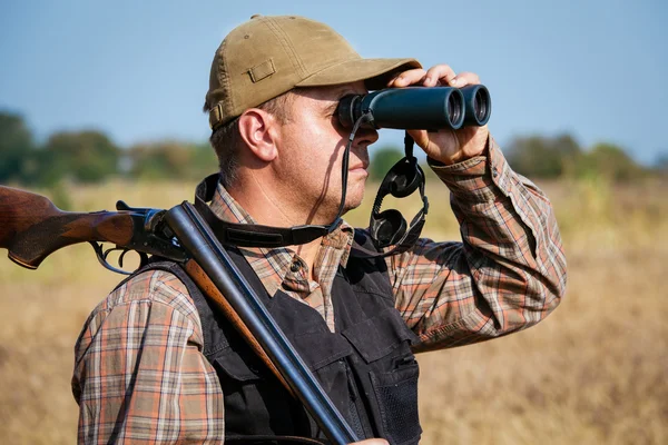 Человек-охотник с дробовиком смотрит через бинокль в лесу — стоковое фото