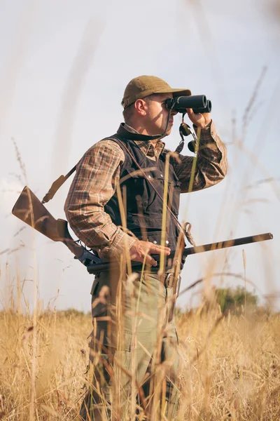 Homem caçador com espingarda olhando através de binóculos na floresta — Fotografia de Stock