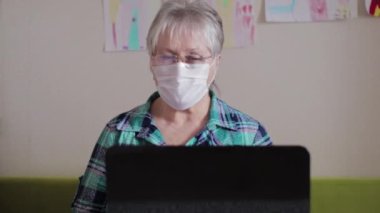 Laptopta koruyucu tıbbi maske takan yaşlı bir kadın. Karantina sırasında internette konuşan bir kadın.