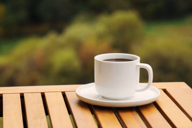 Beyaz bir kahve fincanı altında bir fincan tabağı olan ahşap bir masanın üzerinde oturur. Bardak koyu kahveyle dolu ve sahne arka planda ağaçlarla dolu doğal bir ortamda geçiyor.