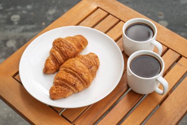 Bir tabak kruvasan ve iki fincan kahve ahşap bir masada oturur. Kruvasanlar altın kahverengidir ve yeni pişirilmiş gibi görünüyor. Kahve fincanları koyu sıvıyla dolu.