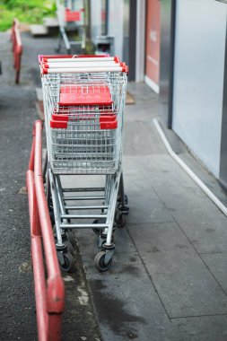 Kaldırımda kırmızı metal korkulukların yanına bir alışveriş arabası park edilmiş. Araba boş ve kırmızı saplı.