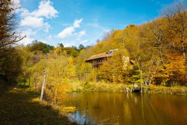 Bir ev bir göl kenarındaki yamaçta bulunur. Ev ağaçlarla çevrili ve göl sakin ve huzurlu. Gökyüzü açık ve güneş ışıl ışıl parlıyor. Sahne huzurlu ve resimli.