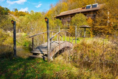 Arka planda bir ev olan derenin üzerinde tahta bir köprü. Köprü eski ve kırsal bir havası var. Ev önünde bir bahçe olan küçük bir kulübedir. Huzurlu ve huzurlu bir sahne.