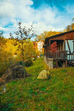 Önünde verandası ve ağacı olan bir ev. Ev bir yaprak yığını ve büyük bir kayayla çevrili. Gökyüzü mavi ve güneş parlıyor.