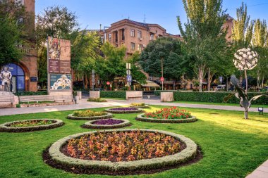 Arkasında büyük bir bina olan bir park. Park çiçekler ve ağaçlarla dolu. Çiçekler dairesel bir şekildedir ve farklı renklerdedir. Park huzurlu bir yer.