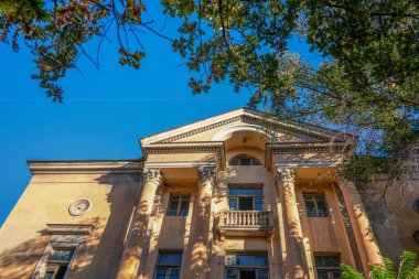 Bir sürü penceresi ve balkonu olan büyük bir bina. Bina kahverengi ve çok detay içeriyor. Gökyüzü mavi ve güneş parlıyor.