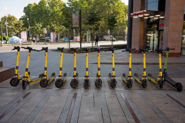 Erivan, Ermenistan 10.16.2025. Bir sıra sarı scooter kaldırıma dizilmiş. Mobiletler yan yana park edilmiş ve birkaç kişi buradan geçiyor. Kentsel yaşam ve ulaşım kavramı