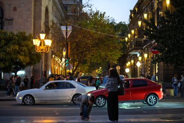 Erivan, Ermenistan 10.16.2025. Kaldırımda kırmızı bir arabanın yanında bir kadın duruyor. Sokaklar insanlarla ve arabalarla dolu.