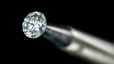 Mücevher cımbızlı elmasın yakın çekimi. Elmas görüntünün ana odak noktasıdır ve metal nesne de elması tutmak ve incelemek için kullanılan stilize bir araçtır.