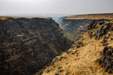 İçinden nehir akan kayalık bir yamaç. Gökyüzü bulutlu ve çimenler kuru.