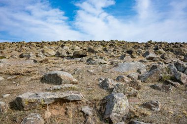 Açık mavi gökyüzü olan kayalık bir yamaç. Gökyüzü bulutlarla kaplı ve kayalar tepenin dört bir yanına dağılmış. Sahne huzurlu ve sakin, kayalar istikrar hissi veriyor.