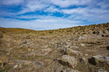 Açık mavi gökyüzü olan kayalık bir yamaç. Gökyüzü bulutlarla kaplı ve yamaçta kayalar var. Sahne huzurlu ve sakin, kayalar istikrar hissi veriyor.