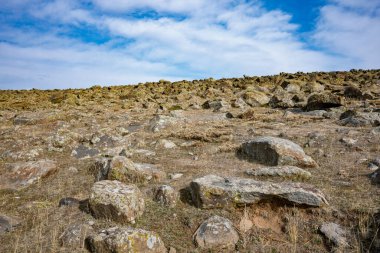 Açık mavi gökyüzü olan kayalık bir yamaç. Gökyüzü bulutlarla kaplı ve kayalar tepenin dört bir yanına dağılmış. Sahne huzurlu ve sakin, kayalar istikrar hissi veriyor.