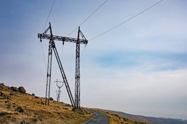 Uzun bir elektrik hattı kulesi sola doğru eğiliyor. Gökyüzü mavi ve bazı bulutlar var.