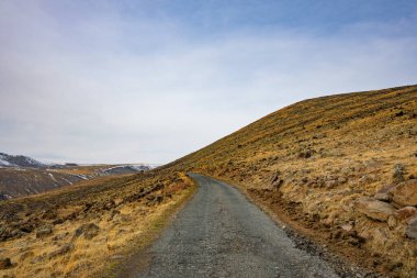 Tepenin yukarısında açık mavi gökyüzü olan bir yol var. Yol çakıl ve kayalardan yapılmıştır ve yamaç kuru otlarla kaplıdır. Huzurlu ve huzurlu bir sahne.