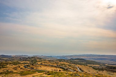 Arka planda dağ sırası olan bulutlu bir gökyüzü. Gökyüzü çoğunlukla mavi ve bazı beyaz bulutlar etrafa saçılmış. Dağ sırası yeşil ve kahverengiyle kayalık arazinin karışımıdır.