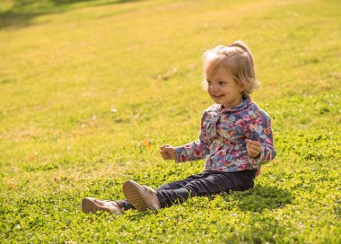 Küçük bir kız çimlerin üzerinde oturuyor ve elinde bir çiçek tutuyor. Sahne huzurlu ve huzurlu, kızın açık havanın ve doğanın güzelliğinin tadını çıkardığı