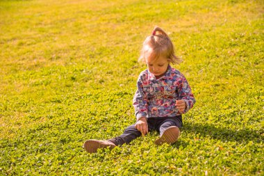 Küçük bir kız çimlerin üzerinde oturuyor ve bir şeye bakıyor. Çiçekli gömlek ve kot pantolon giyiyor.