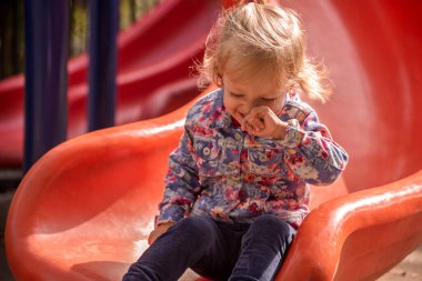 Küçük bir kız kırmızı kaydırağın üzerinde oturuyor, ağlıyor. Mavi bir ceket ve kot pantolon giyiyor.