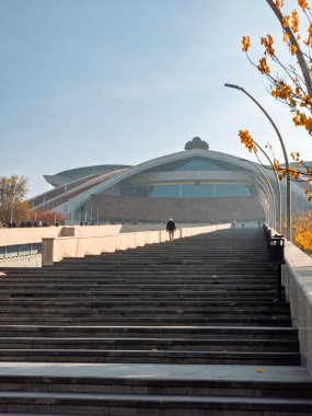 Karen Demirtchian 'dan sonra Sport Concert Complex' in büyük binasının önünde merdivenlerden çıkan bir kişi var. Bina yüksek ve bir sürü penceresi var. Gökyüzü açık ve güneş parlıyor.