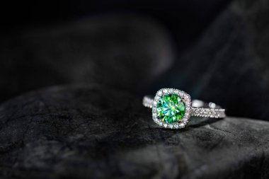 Siyah bir taşın üzerinde yeşil bir elmas yüzük duruyor. Taş karanlık ve yüzük parlak.